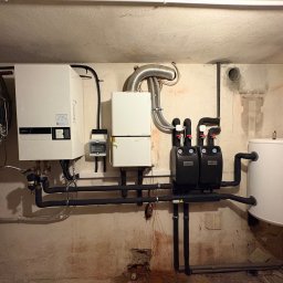 Dawid Tomaszewski - Złożona instalacja grzewcza w piwnicy: białe kotły, czarne rury, pompy obiegowe Termojet, zbiornik buforowy i rower oparty o ścianę. Widoczne ślady wilgoci na ścianach.