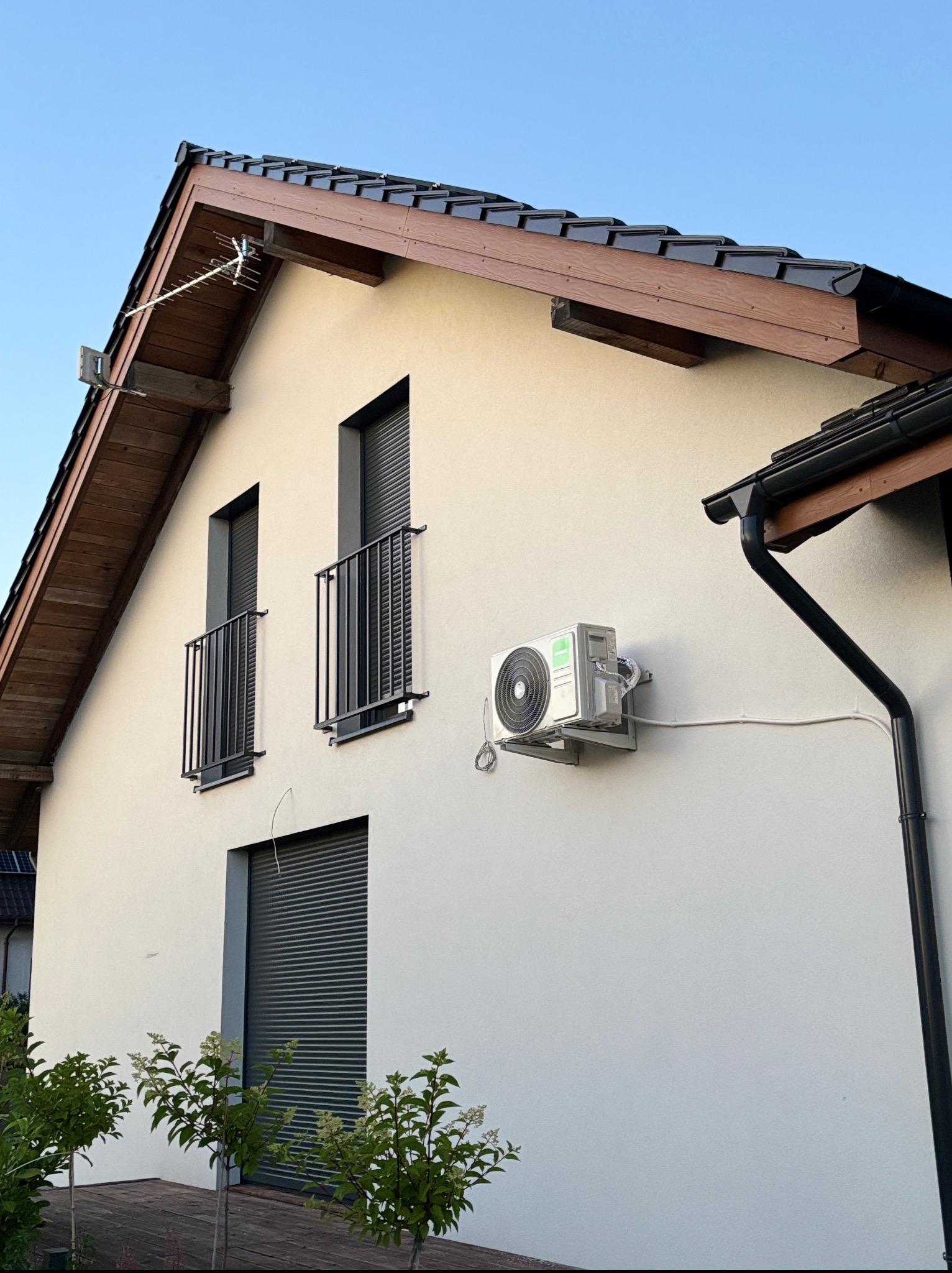 Zewnętrzna jednostka klimatyzacji KAISAI na elewacji domu z oknami balkonowymi i brązową dachówką. Widoczna antena na dachu i rynna. Biała ściana, błękitne niebo.
