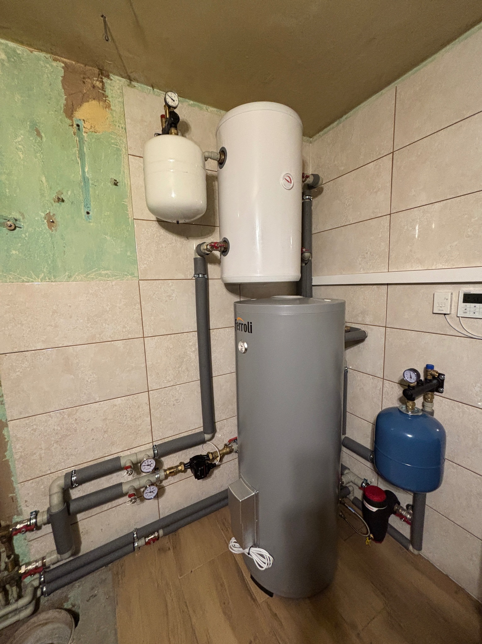 Szara pompa ciepła Ferroli z białym zasobnikiem c.w.u. i niebieskim zbiornikiem wyrównawczym. Widoczne rury i manometry. Część ściany pomalowana na zielono.