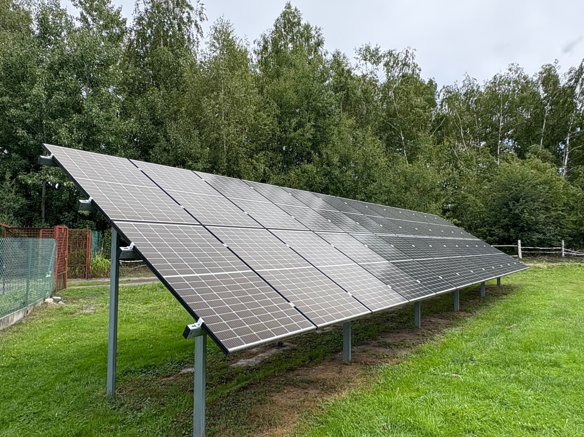 Panel fotowoltaiczny na metalowej konstrukcji, otoczony zielenią. Widoczne drzewa w tle. Instalacja na trawiastym terenie, częściowo ogrodzona siatką.