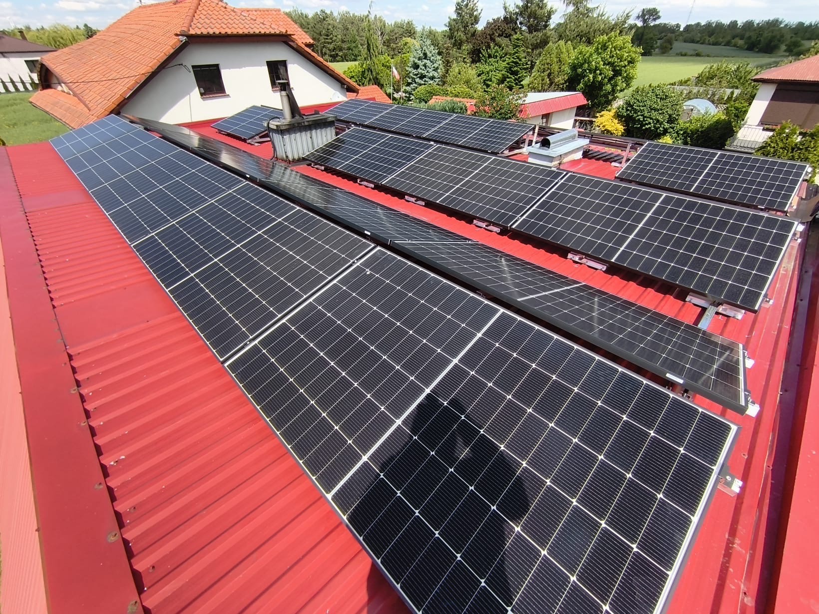 Panele fotowoltaiczne na czerwonym dachu domu jednorodzinnego, widok z góry. Moduły ułożone rzędami, instalacja zakończona. W tle zieleń drzew i budynki.