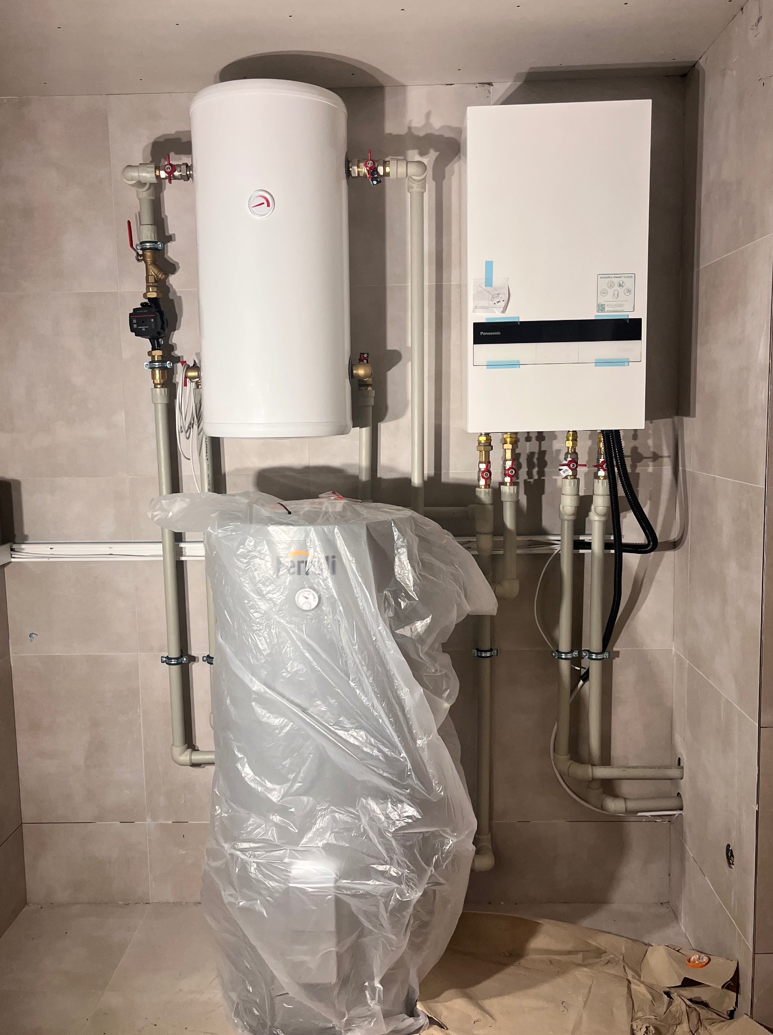 Biały zasobnik ciepłej wody i panel sterowania Panasonic na ścianie z szarych płytek, rury z zaworami. Dolny zasobnik w folii ochronnej.