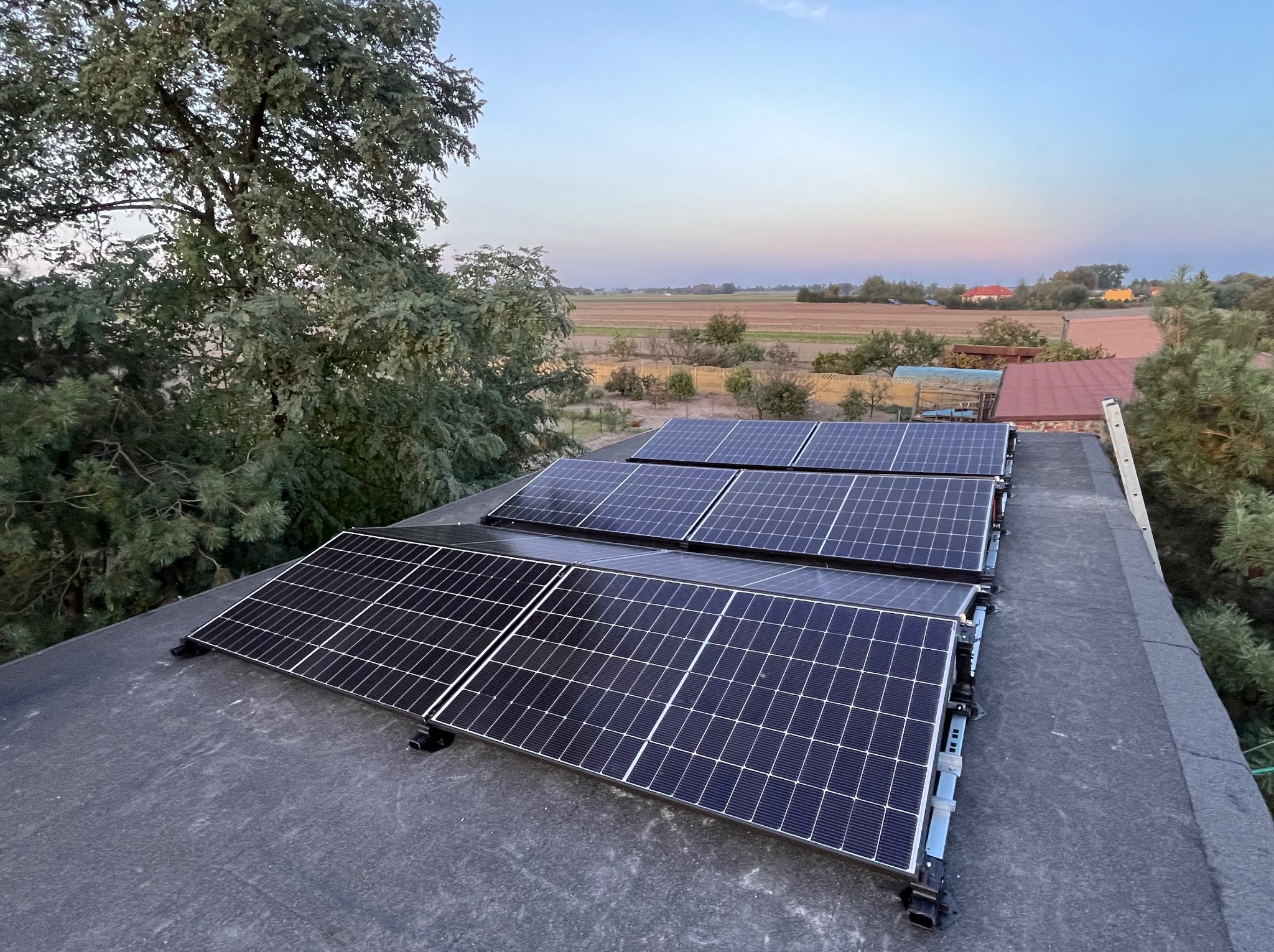 Panele fotowoltaiczne na dachu, widok z góry na tle zieleni i pól uprawnych o zmierzchu. Układ paneli w rzędach, widoczne mocowania i fragment drabiny.