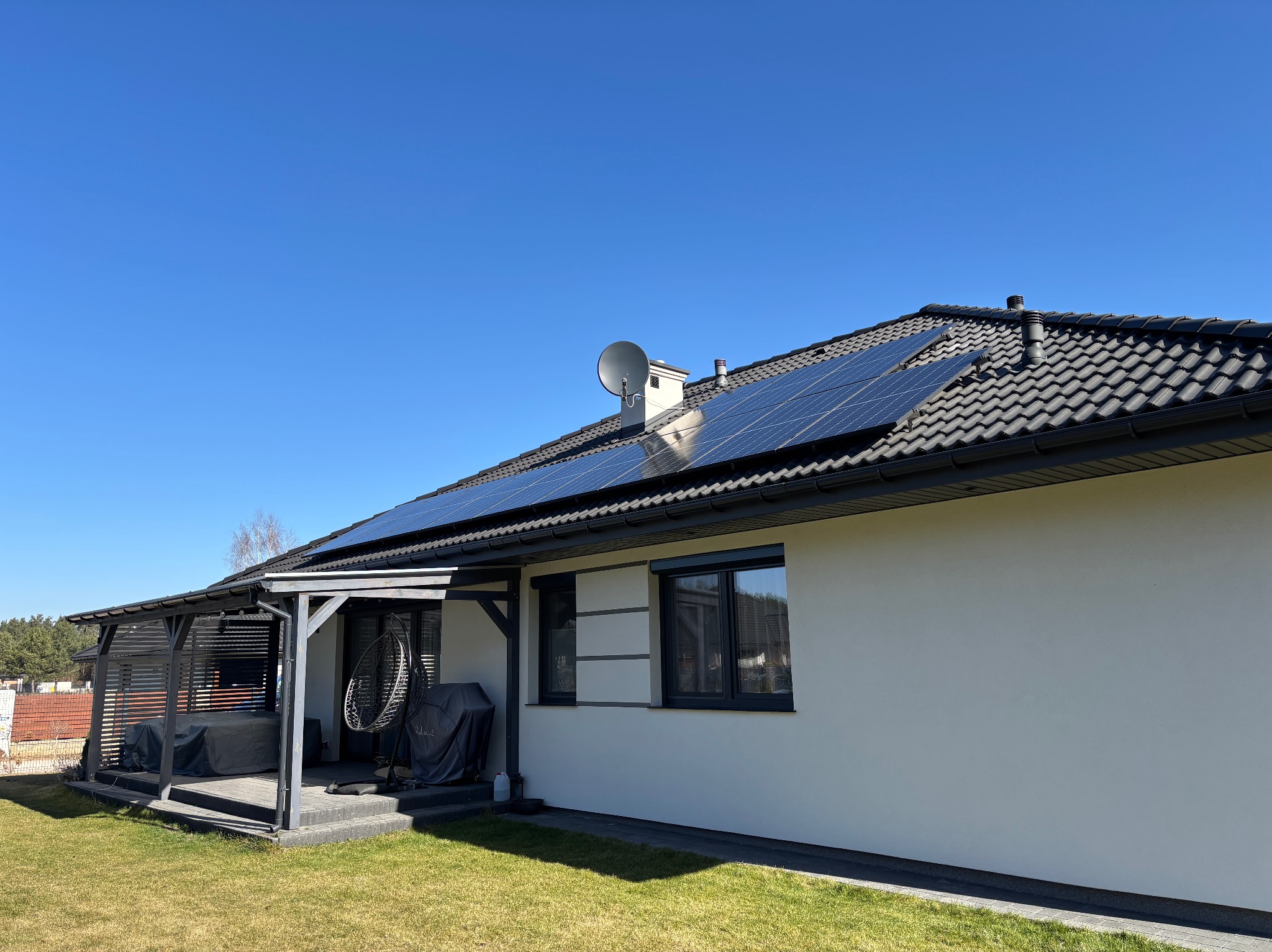 Dom jednorodzinny z panelami fotowoltaicznymi na dachu, widok z ogrodu w słoneczny dzień. Na dachu widoczna antena satelitarna.