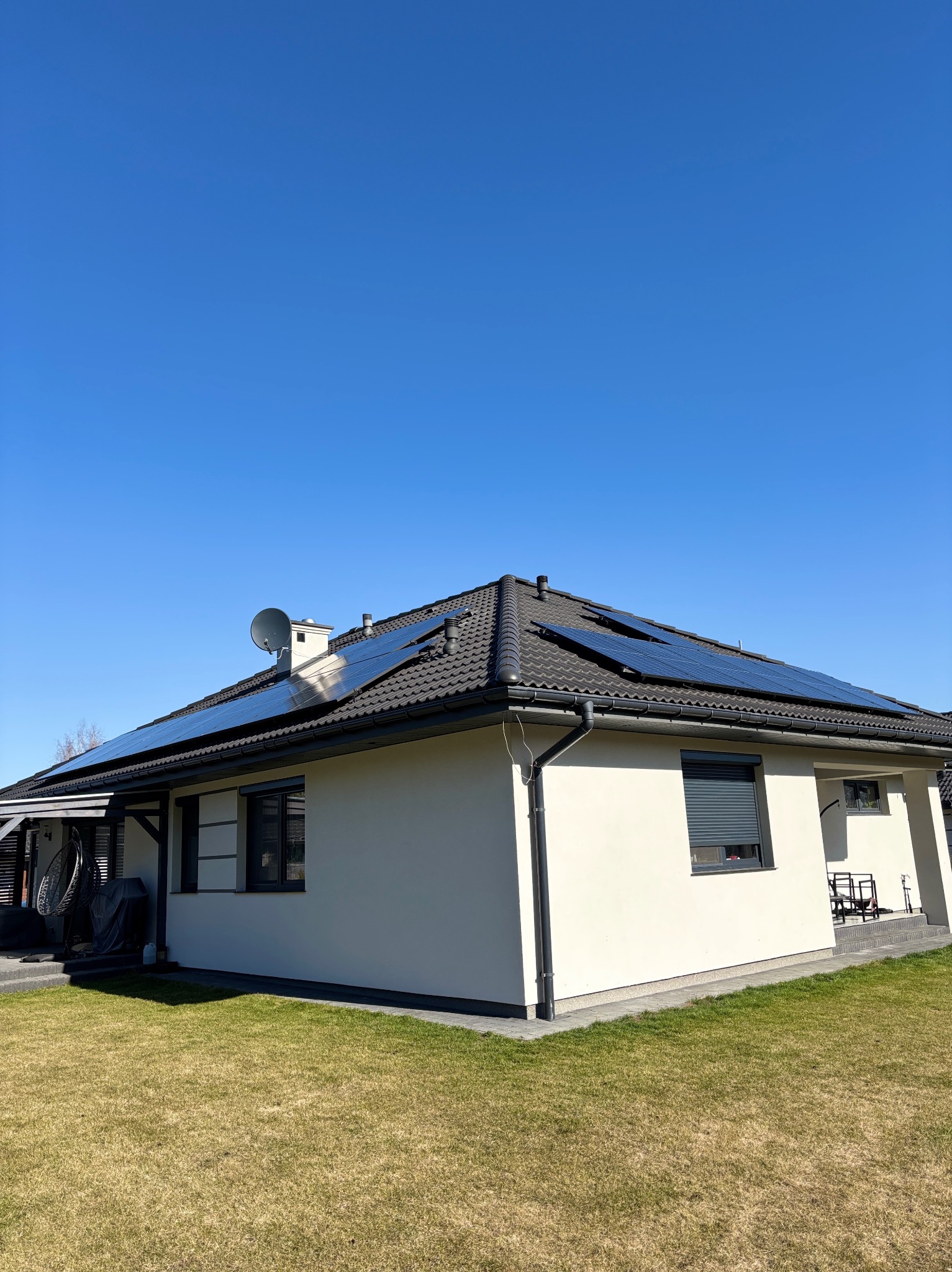 Dom jednorodzinny z panelami fotowoltaicznymi na dachu w słoneczny dzień. Widoczna antena satelitarna i trawnik przed budynkiem. Minimalistyczna architektura.