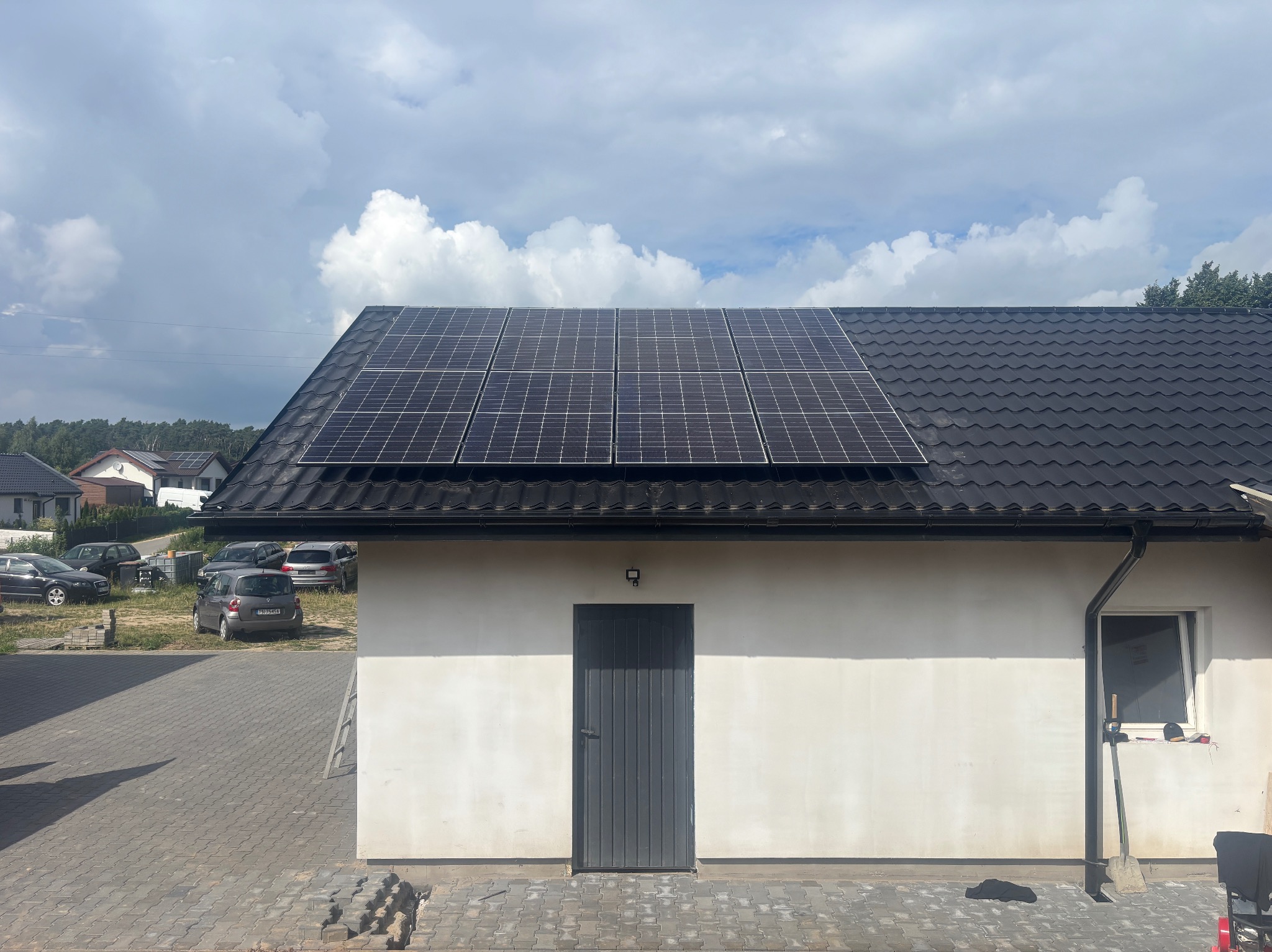 Panele słoneczne na ciemnym dachu budynku z białą elewacją. W tle błękitne niebo z chmurami. Na pierwszym planie kostka brukowa i fragment posesji.