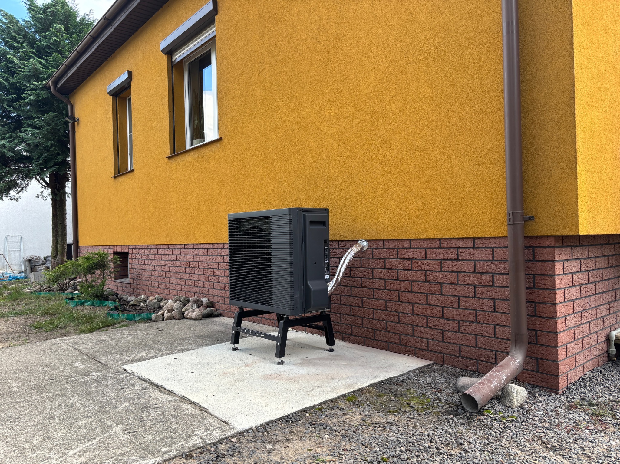 Czarna pompa ciepła na betonowej podstawie, obok żółtego budynku z brązową cegłą i rynną. Widoczne okna z roletami, drzewo i kamienie dekoracyjne.