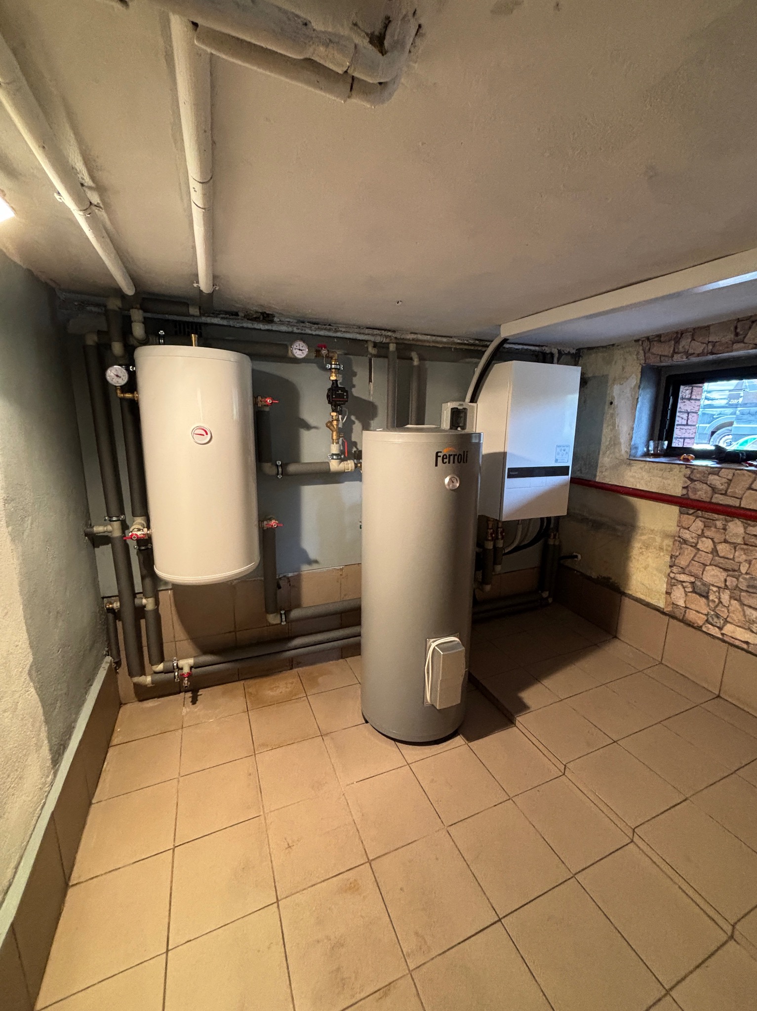 Piwnica z dwoma bojlerami i kotłem gazowym, widoczne rury i armatura. Jeden bojler marki Ferolli. Kamienna ściana z małym oknem w tle.