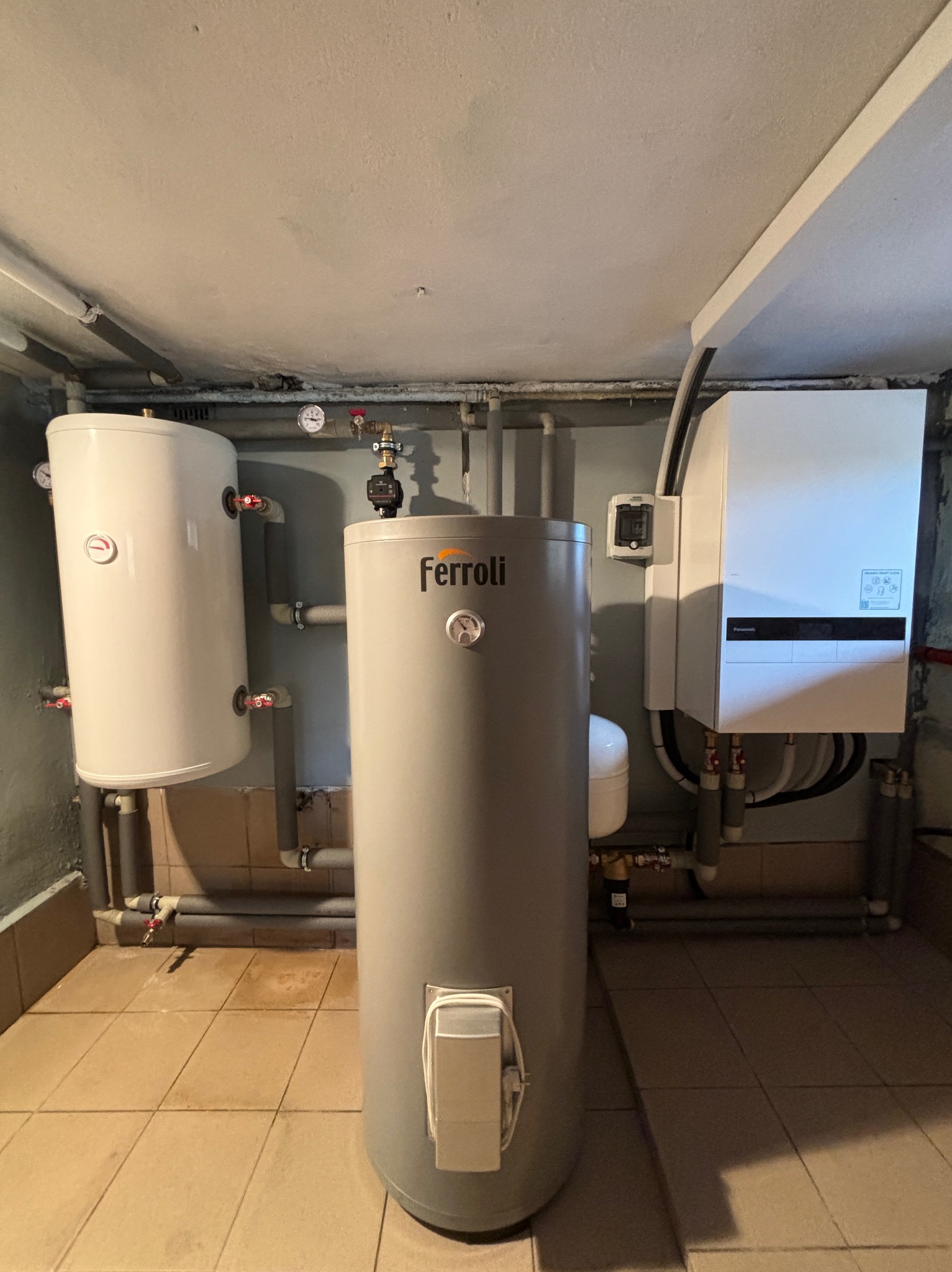 Kompletna instalacja grzewcza w piwnicy: szary zbiornik Ferroli, biały zasobnik CWU i piec gazowy Panasonic. Widoczne rury, zawory i manometry.