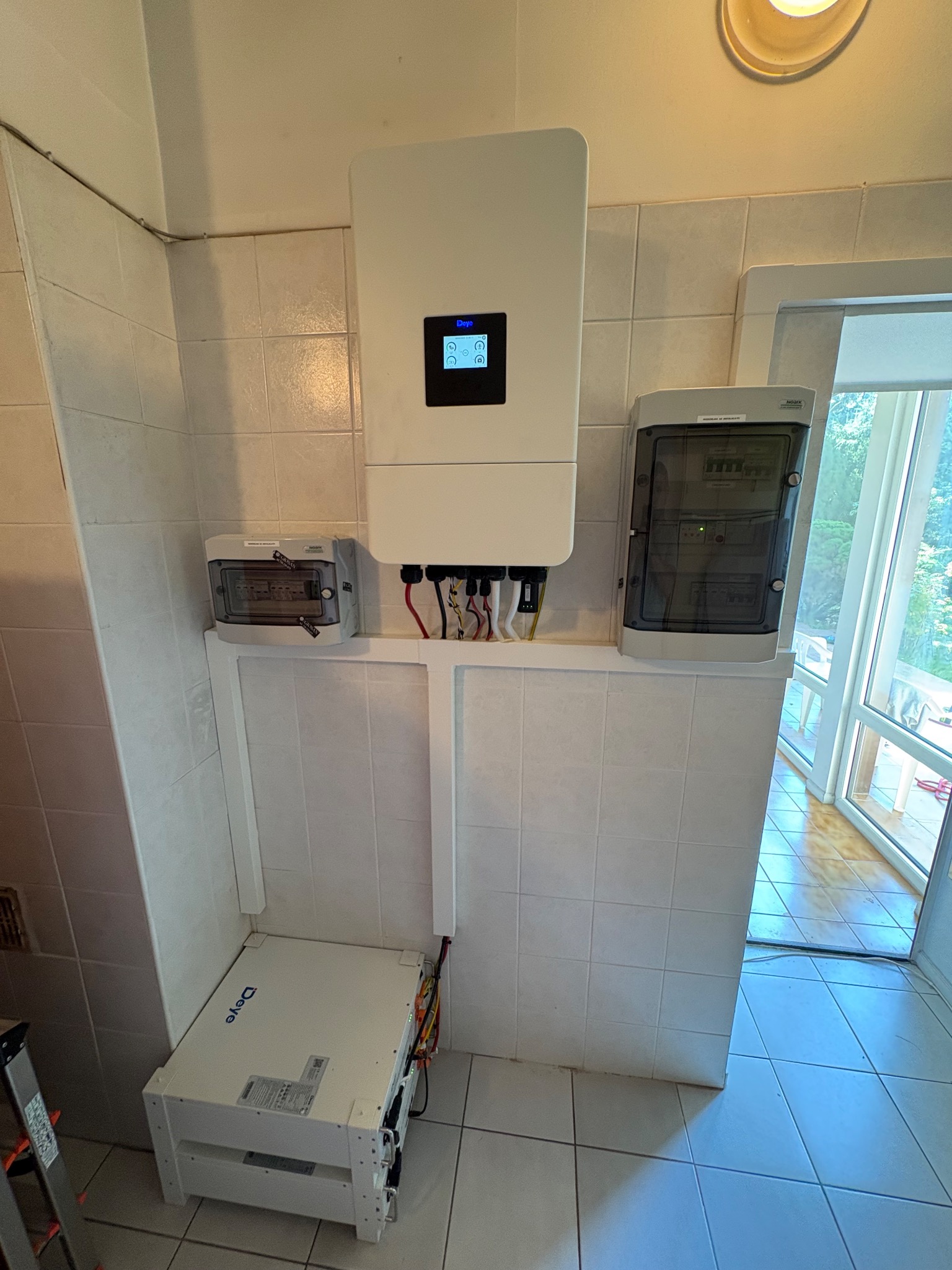 Wnętrze z zamontowanym białym inwerterem i akumulatorem marki Deye na tle kafelkowanej ściany i podłogi. Widoczna skrzynka elektryczna i fragment drabiny.