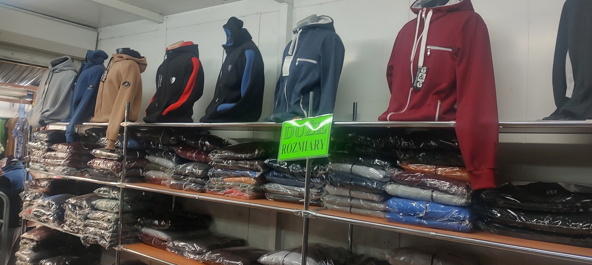 Regał z bluzami z kapturem, zarówno na manekinach, jak i zapakowanymi w folię, z zieloną tabliczką 'DUŻE ROZMIARY' w tle.