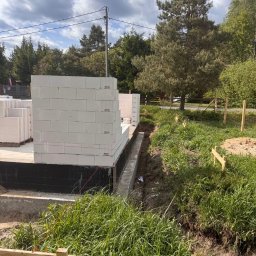 ARBUD Artur Michalak - Budowa domu: fundamenty z izolacją i pierwsze warstwy ścian z białego bloczka, widok z poziomu gruntu, zieleń w tle.