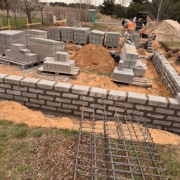 ARBUD Artur Michalak - Budowa fundamentów domu z bloczków betonowych. Widoczne zbrojenie, zapas bloczków na paletach, betoniarka i wykop pod fundament. Prace w toku, pochmurny dzień.