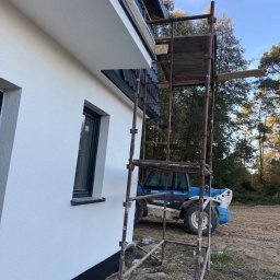 ARBUD Artur Michalak - Ściana budynku z oknem po ociepleniu, widoczne rusztowanie i maszyna budowlana w tle. Elewacja w kolorze białym.