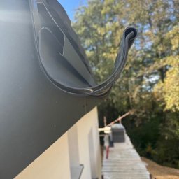 ARBUD Artur Michalak - Zamontowana, czarna rynna dachowa z bliska, na tle fragmentu elewacji domu i rusztowania. Widok z dołu, błękitne niebo i drzewa w tle.