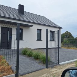 ARBUD Artur Michalak - Nowoczesny dom z ciemnym dachem i szarą elewacją, ogrodzony metalową siatką. Widoczny podjazd wyłożony kostką brukową i fragment zieleni przy wejściu.
