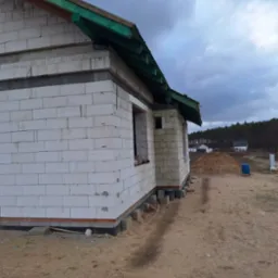 Budowa domu jednorodzinnego w trakcie realizacji, ściany z białych bloczków, widoczna więźba dachowa z zielonymi elementami, otoczenie placu budowy.