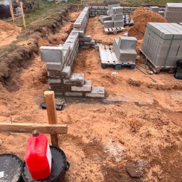 ARBUD Artur Michalak - Budowa fundamentów z bloczków betonowych. Częściowo wymurowane ściany fundamentowe, palety z bloczkami, wykop. Widok z góry, ziemia wokół.