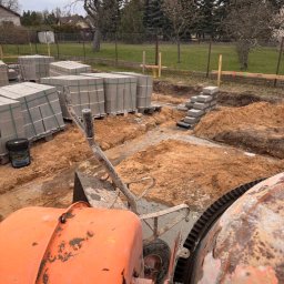 ARBUD Artur Michalak - Fundamenty w trakcie budowy: wykop, bloczki betonowe na paletach, częściowo wymurowana ściana fundamentowa, pomarańczowa betoniarka. Widok na zewnątrz, w tle drzewa i ogrodzenie.