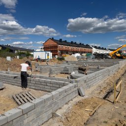 ARBUD Artur Michalak - Wznoszenie fundamentów z bloczków betonowych na placu budowy. Widoczny pracownik, częściowo wymurowane ściany fundamentowe i sprzęt budowlany w tle.