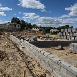 ARBUD Artur Michalak - Wznoszenie murów fundamentowych z bloczków betonowych na budowie. Widoczne palety z materiałem i drewniane szalunki. Błękitne niebo z chmurami w tle.