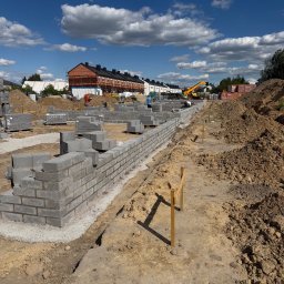 ARBUD Artur Michalak - Wznoszenie muru z bloczków betonowych na fundamencie. Widoczne sznurki murarskie, palety z bloczkami i koparka w tle. Słoneczny dzień, błękitne niebo z chmurami.
