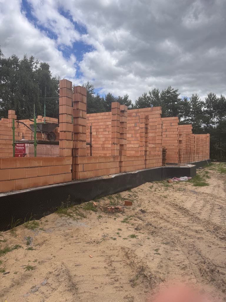 Wznoszenie ścian z czerwonej cegły Porotherm na fundamencie. Widoczne zbrojenia i paleta cegieł. W tle drzewa i pochmurne niebo.