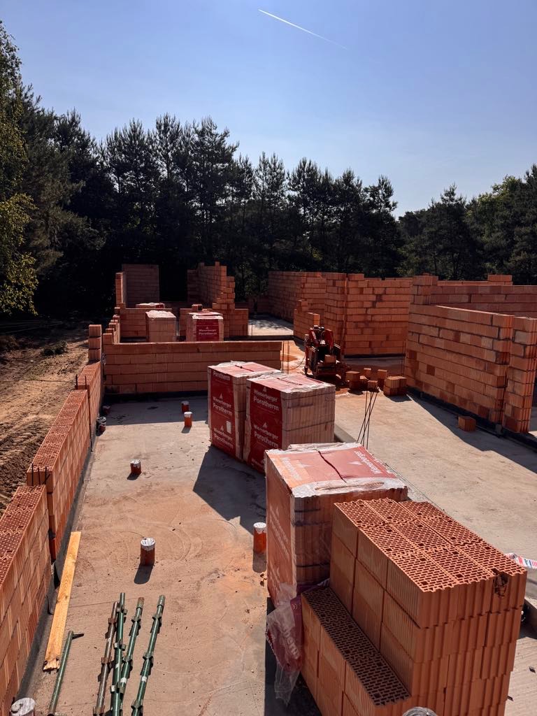 Budowa domu z czerwonej cegły Porotherm. Częściowo wzniesione ściany, palety z cegłami na fundamencie. Widok z góry, las w tle, błękitne niebo.