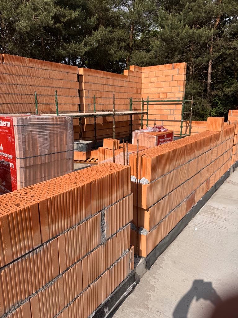 Wznoszenie ścian z czerwonej ceramiki Porotherm na budowie domu. Widoczne rusztowania i paleta z materiałem. Świeżo wymurowane ściany, oświetlone słońcem.