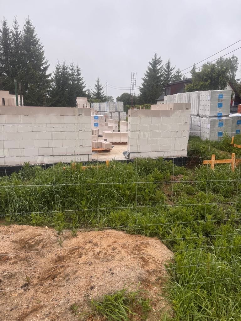 Budowa domu w trakcie: wzniesione ściany z białych bloczków, palety z materiałami w tle, teren budowy otoczony siatką. Widok z poziomu gruntu.