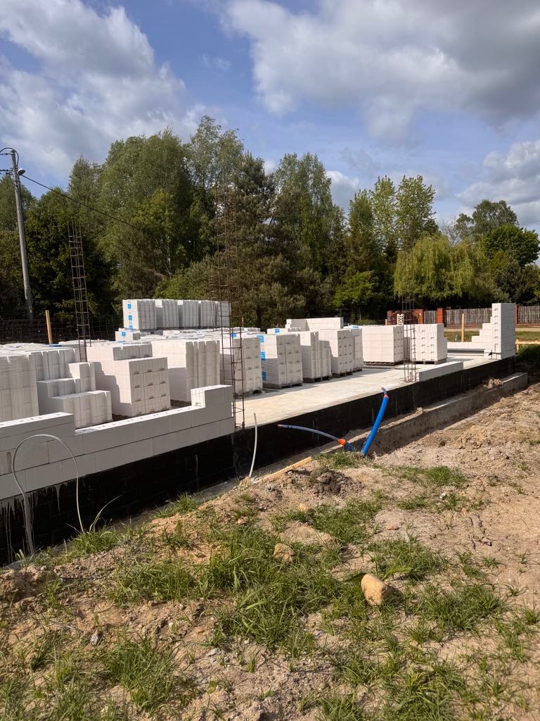 Wznoszenie ścian z białych bloczków na fundamencie z izolacją. Częściowo wymurowane ściany i palety z bloczkami na placu budowy, widok z poziomu gruntu.