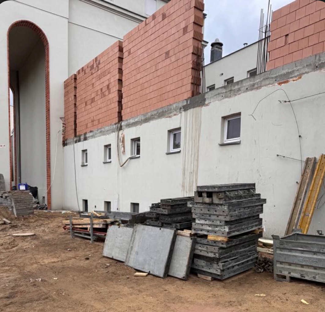 Budowa z cegły: ściana w trakcie murowania na tle jasnej elewacji z oknami. Na pierwszym planie stosy szalunków i materiałów budowlanych na ziemi.