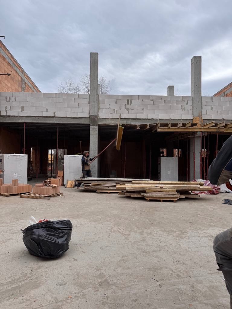 Budowa domu z betonu komórkowego w stanie surowym otwartym, widoczne ściany, słupy, strop, drewniane elementy szalunku i pracownik z narzędziem.