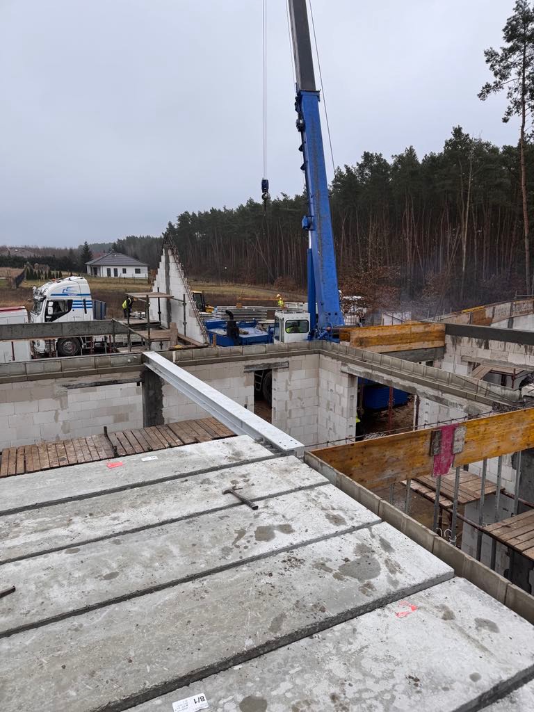 Budowa domu z pustaków: widok z góry na strop z płyt prefabrykowanych, dźwigiem transportowane są kolejne elementy. W tle las i fragment budynku.