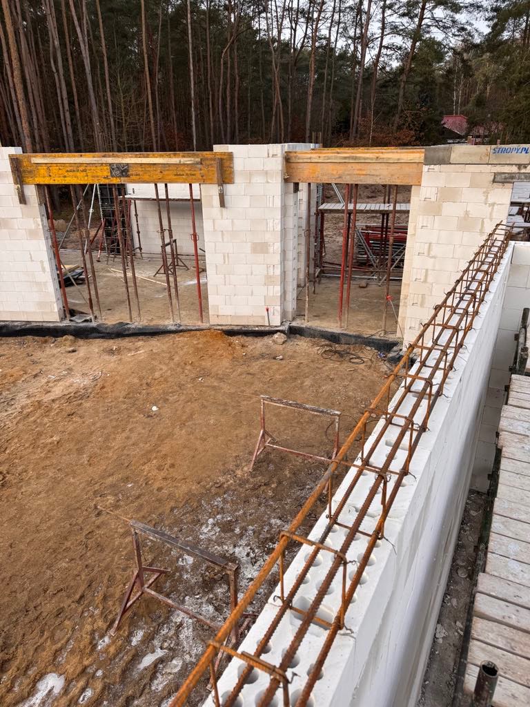 Wznoszone ściany z betonu komórkowego, widoczne zbrojenia, szalunki nad otworami okiennymi. Prace budowlane w toku, widok z góry na fundamenty domu w lesie.