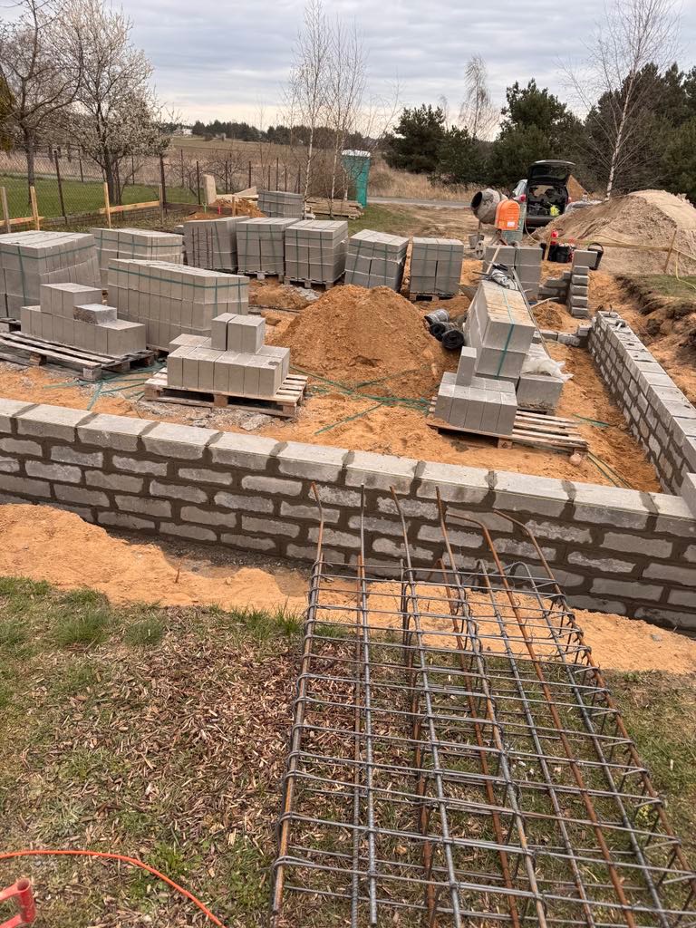 Budowa fundamentów domu z bloczków betonowych. Widoczne zbrojenie, zapas bloczków na paletach, betoniarka i wykop pod fundament. Prace w toku, pochmurny dzień.