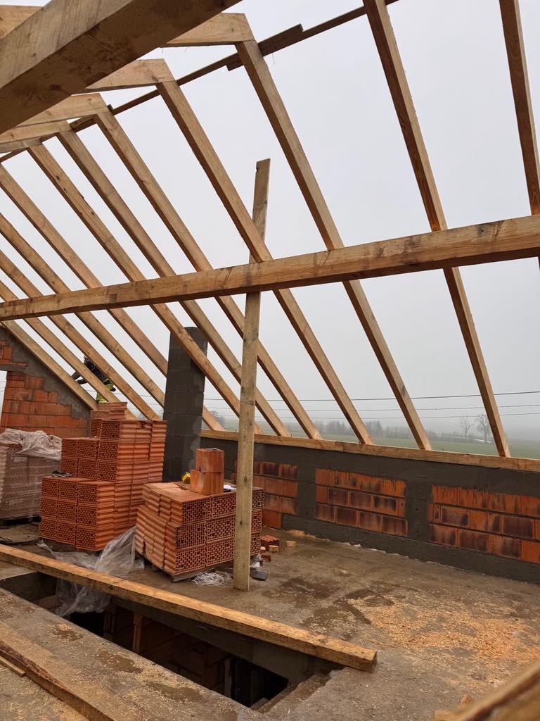 Wnętrze w trakcie budowy: surowa więźba dachowa z drewnianych belek, częściowo wymurowane ściany z czerwonej cegły i palety cegieł. Szary, pochmurny dzień w tle.