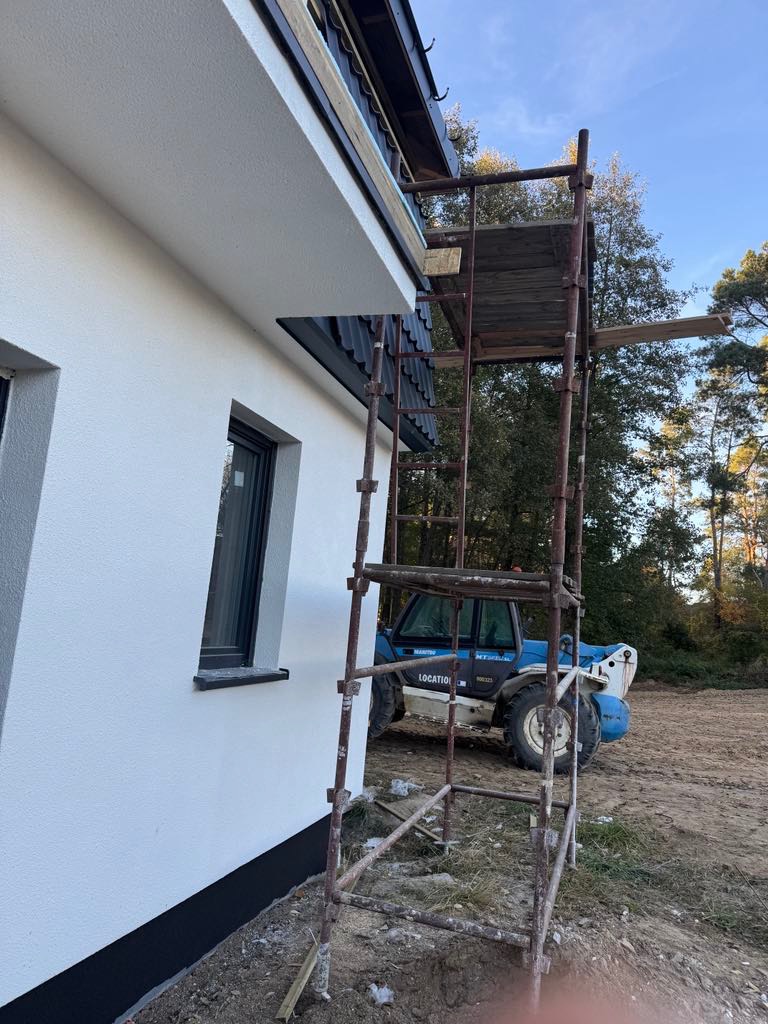 Ściana budynku z oknem po ociepleniu, widoczne rusztowanie i maszyna budowlana w tle. Elewacja w kolorze białym.