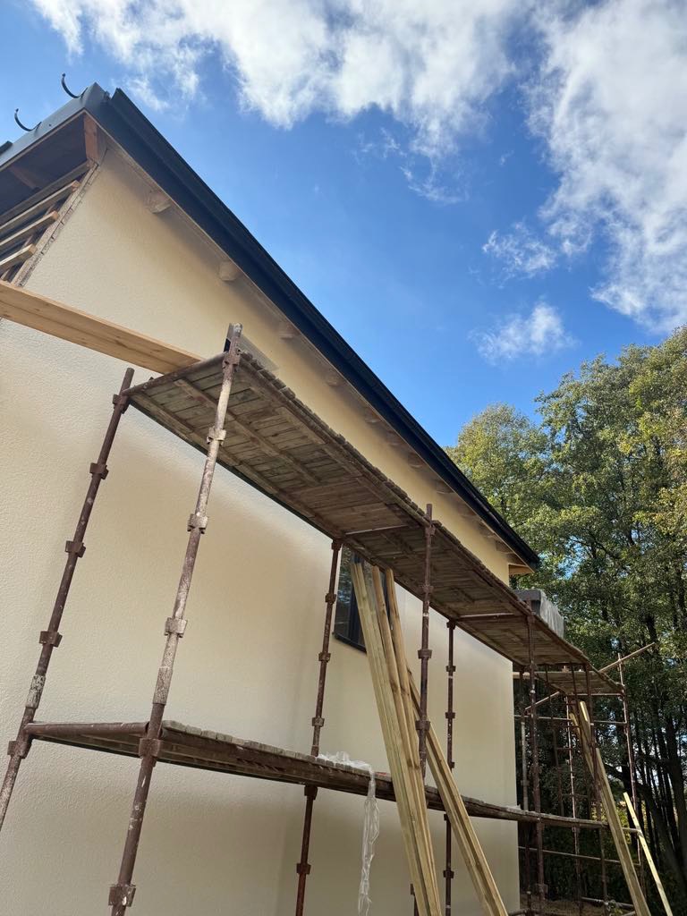 Ocieplony dom z elewacją w kolorze kremowym, widok z rusztowania. Świeża elewacja, błękitne niebo z chmurami w tle. Prace wykończeniowe.