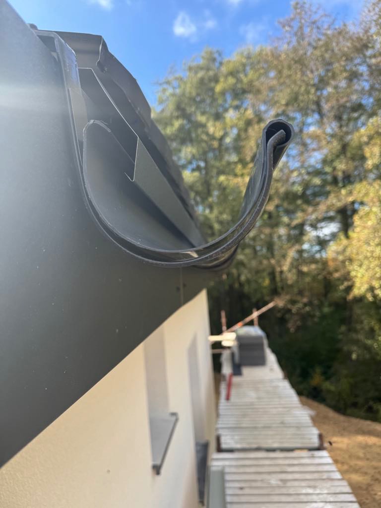 Zamontowana, czarna rynna dachowa z bliska, na tle fragmentu elewacji domu i rusztowania. Widok z dołu, błękitne niebo i drzewa w tle.