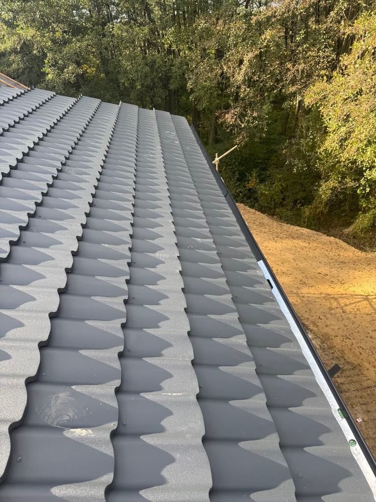 Szary dach z blachy trapezowej, widok z góry, z poziomnicą w rogu, na tle drzew. Widoczne mocowania blachy. Krycie dachu w trakcie realizacji.