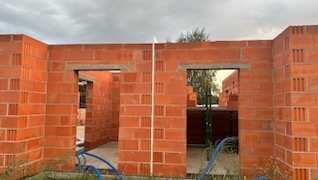 Czerwone, porothermowe ściany domu w trakcie budowy. Widoczne otwory okienne i drzwiowe, szare nadproża. W tle zieleń i pochmurne niebo.