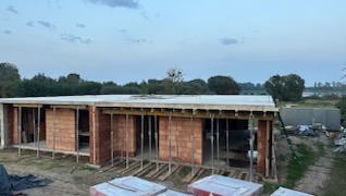 Niski budynek w trakcie budowy z czerwonej cegły, z widoczną konstrukcją dachu i podporami. Na pierwszym planie paczki cegieł. Tło: zieleń, błękitne niebo.