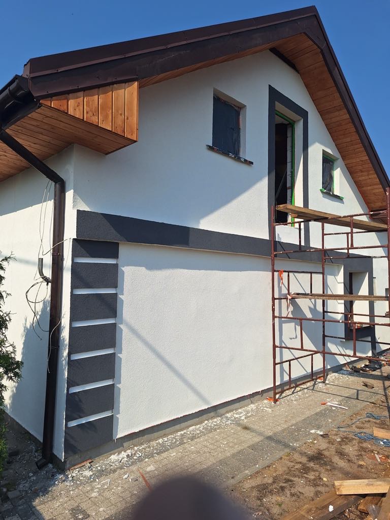 Ocieplony dom z białą elewacją, dekoracyjnymi szarymi elementami i drewnianą podbitką. Widoczne rusztowanie i zabezpieczone okna. Słoneczny dzień, boczny kadr.