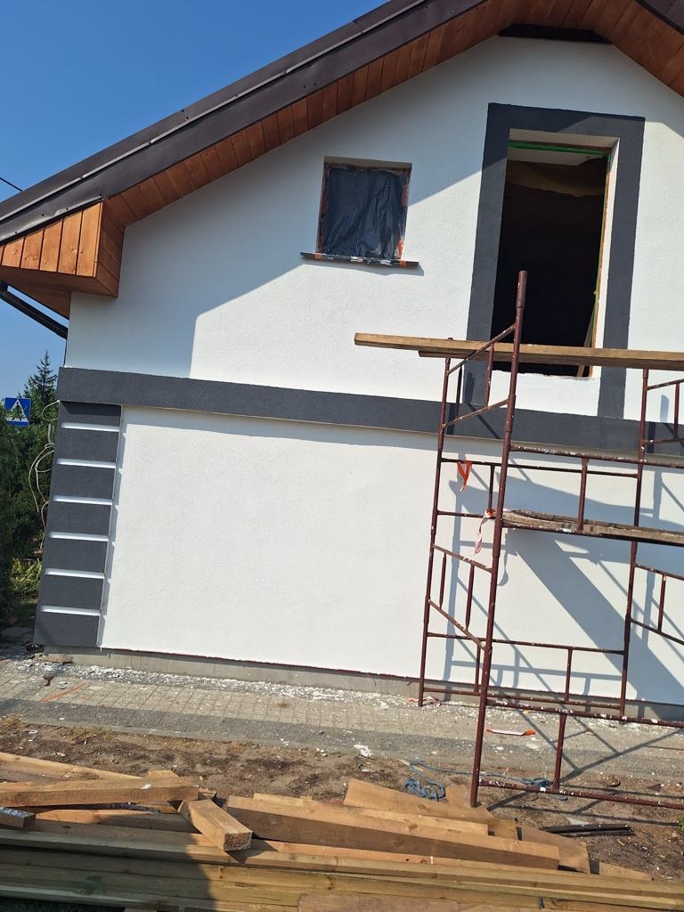 Boczna ściana domu w trakcie ocieplania: biały tynk, szara listwa, okno zabezpieczone folią, rusztowanie i drewno. Prace budowlane w toku.