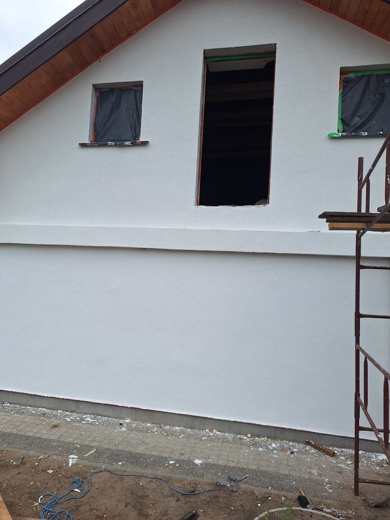 Biała fasada domu w trakcie budowy, okna zabezpieczone folią, widoczne rusztowanie, drewniany dach, surowy teren wokół budynku.