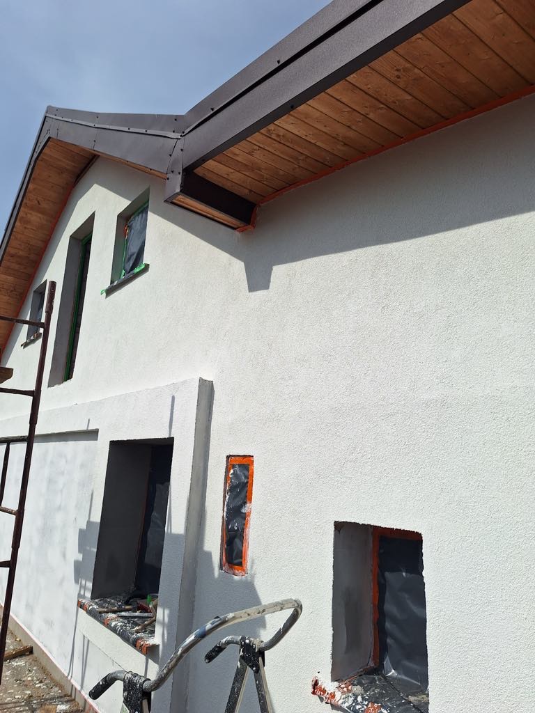 Biała ściana domu w trakcie prac wykończeniowych z widocznymi oknami zabezpieczonymi folią, drewnianą podbitką i fragmentem rusztowania.