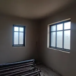 Pokój po tynkowaniu maszynowym: dwie okna z czarnymi ramami, szare ściany, stos rusztowań leżący na podłodze. Widok na świeżo otynkowane ściany.