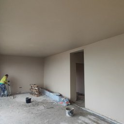 Bizland - Wnętrze po tynkowaniu maszynowym: gładkie ściany w beżowym odcieniu, duże okno z widokiem na zewnątrz, pracownik z narzędziem na podłodze. Widoczne ślady prac.