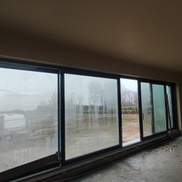 Bizland - Wnętrze w trakcie prac wykończeniowych: pracownik szpachluje ścianę. Duże okno z widokiem na zewnątrz, na podłodze resztki materiałów budowlanych, widoczne ślady szpachlowania.